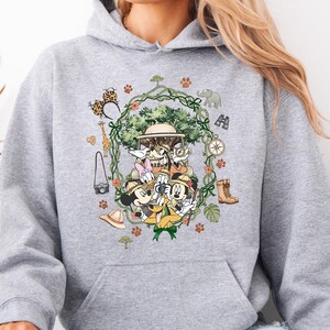 Peut inclure: Un sweat &agrave; capuche gris clair avec un motif color&eacute; de personnages de dessins anim&eacute;s en tenue de safari. Le motif comprend un arbre, des animaux et des accessoires sur le th&egrave;me du safari. Le sweat &agrave; capuche a une poche kangourou et une capuche &agrave; cordon.