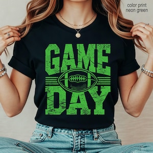 May include: A black short-sleeved t-shirt featuring a neon green graphic print with the text "GAME DAY" and a football illustration. The design has a distressed, textured appearance. The shirt is paired with blue denim jeans.