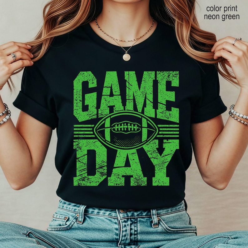 May include: A black short-sleeved t-shirt featuring a neon green graphic print with the text "GAME DAY" and a football illustration. The design has a distressed, textured appearance. The shirt is paired with blue denim jeans.