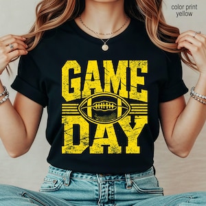 May include: A black short-sleeved t-shirt featuring a distressed yellow graphic print. The design displays the text "GAME DAY" in large block letters with a stylized football illustration positioned horizontally between the two words.