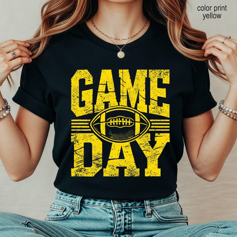 May include: A black short-sleeved t-shirt featuring a distressed yellow graphic print. The design displays the text "GAME DAY" in large block letters with a stylized football illustration positioned horizontally between the two words.
