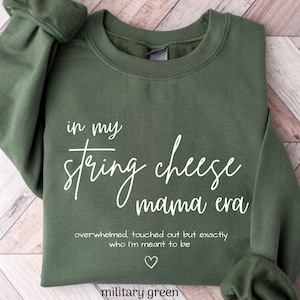 May include: Military green sweatshirt with the text "in my string cheese mama era". The text continues with "overwhelmed, touched out but exactly who I'm meant to be" and a heart symbol. The sweatshirt is a solid color.