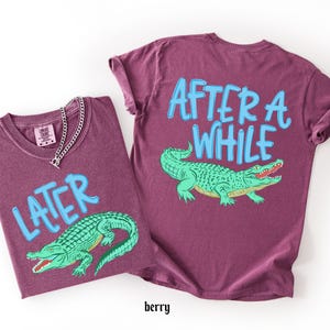 May include: Two berry-colored t-shirts with the text "LATER" and "AFTER A WHILE" in light blue. Each shirt features a green alligator graphic. A silver chain necklace is on one shirt.