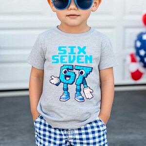 May include: A gray t-shirt with the words "SIX SEVEN" in light blue pixelated font. Below, the numbers 6 and 7 are depicted as cartoon characters with blue bodies, white gloves, and blue shoes. The shirt is worn by a child.