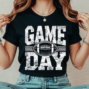 Puede incluir: Camiseta negra con la frase "GAME DAY" en letras blancas desgastadas. Un gr&aacute;fico de f&uacute;tbol americano blanco est&aacute; centrado entre las palabras. La camiseta es de estilo casual.