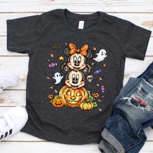 Puede incluir: Camiseta gris oscuro con un dise&ntilde;o de Halloween. El dise&ntilde;o presenta a los personajes de Mickey y Minnie Mouse apilados sobre una calabaza tallada, rodeados de fantasmas, murci&eacute;lagos y caramelos. La camiseta se muestra con vaqueros azules y zapatillas blancas.