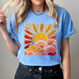 May include: Light blue t-shirt featuring an embroidered design of a sun with orange and yellow rays, and layered clouds in shades of orange and red. The shirt has short sleeves and a crew neck.
