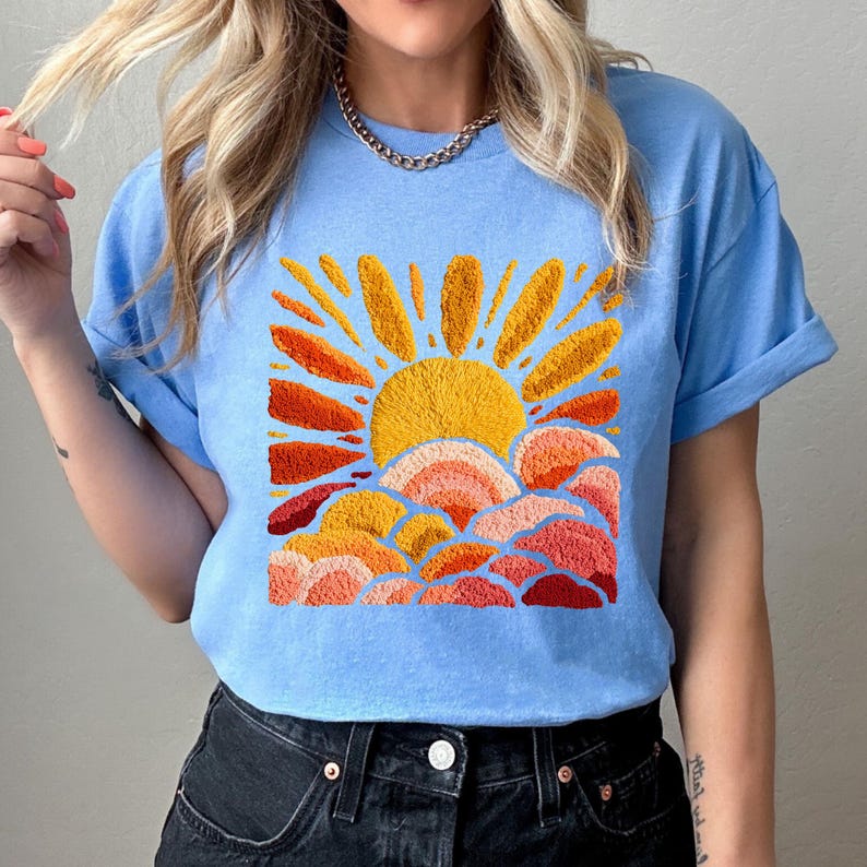 May include: Light blue t-shirt featuring an embroidered design of a sun with orange and yellow rays, and layered clouds in shades of orange and red. The shirt has short sleeves and a crew neck.