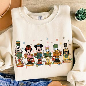 May include: Cream-colored sweatshirt featuring a festive design of Disney characters dressed as nutcrackers. The characters include Mickey Mouse, Minnie Mouse, Donald Duck, and Goofy, each holding a gift or weapon. Decorative stars and snowflakes are also present.