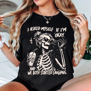 May include: Black t-shirt with a skeleton graphic wearing sunglasses and holding a beverage. The text on the shirt reads, "I asked myself if I'm okay, and we both started laughing."