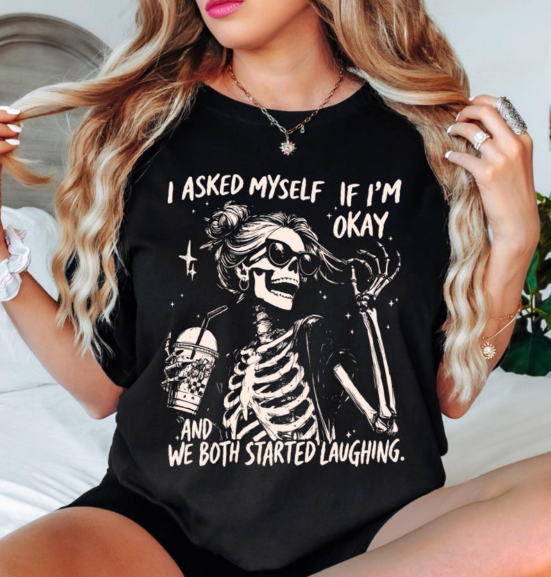 May include: Black t-shirt with a skeleton graphic wearing sunglasses and holding a beverage. The text on the shirt reads, "I asked myself if I'm okay, and we both started laughing."