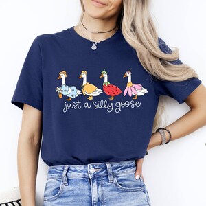 May include: Navy blue t-shirt featuring a whimsical design of four geese dressed in various outfits, including a dress, cape, strawberry, and flower skirt. The text "just a silly goose" is printed below the geese.