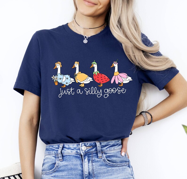May include: Navy blue t-shirt featuring a whimsical design of four geese dressed in various outfits, including a dress, cape, strawberry, and flower skirt. The text "just a silly goose" is printed below the geese.