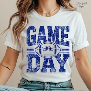 May include: A white short-sleeve t-shirt featuring a distressed royal blue graphic print. The design displays the text GAME DAY in large block letters with a football illustration centered between the words. The text color print royal is visible.