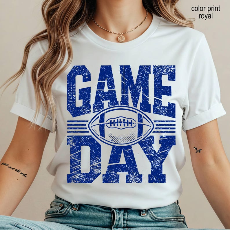 May include: A white short-sleeve t-shirt featuring a distressed royal blue graphic print. The design displays the text GAME DAY in large block letters with a football illustration centered between the words. The text color print royal is visible.