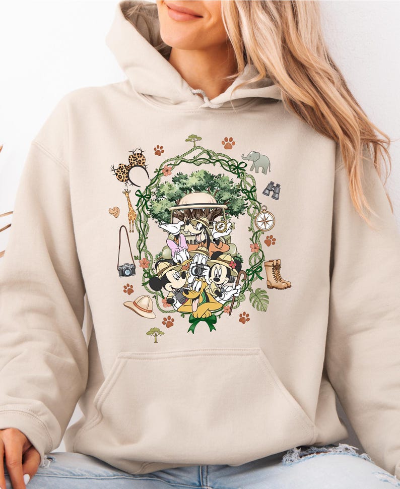 Peut inclure: Sweat &agrave; capuche beige avec un motif color&eacute; de Mickey, Donald, Dingo et Daisy en tenue de safari. Le design comprend des &eacute;l&eacute;ments de safari comme une boussole, des jumelles et une girafe, entour&eacute;s d'une guirlande de vigne verte.