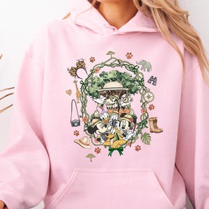 Peut inclure: Un sweat &agrave; capuche rose clair avec un motif de Mickey, Minnie, Donald et Dingo en tenue de safari. Le motif comprend un arbre, des accessoires de safari et des empreintes de pattes. Le sweat &agrave; capuche a une poche avant.