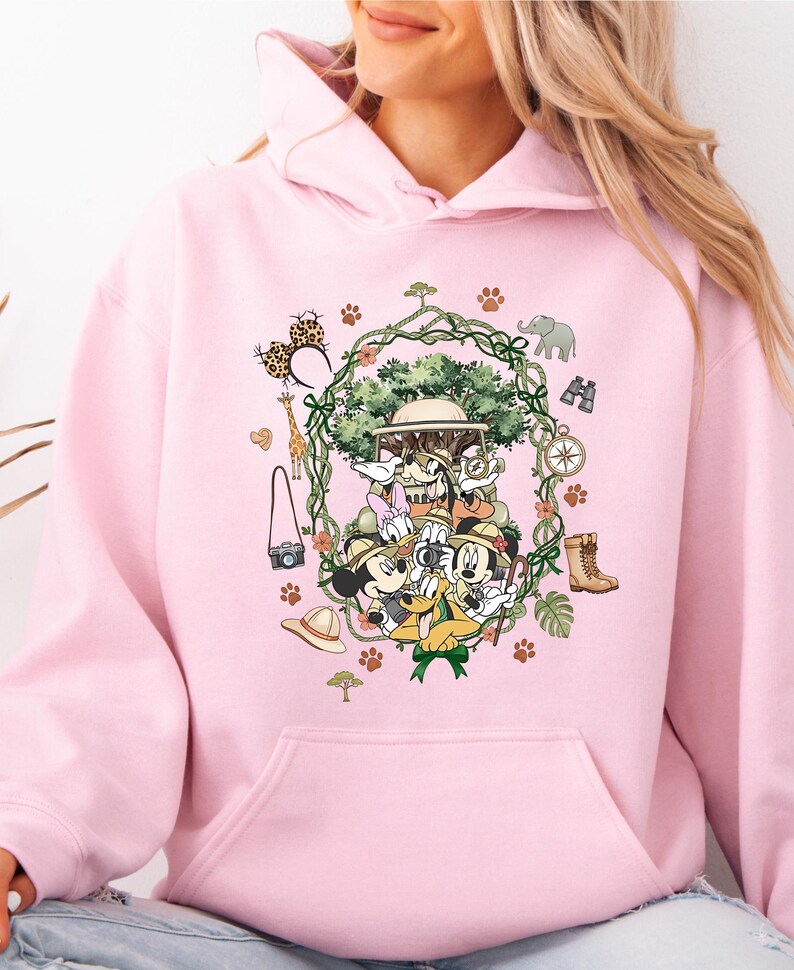 Peut inclure: Un sweat &agrave; capuche rose clair avec un motif de Mickey, Minnie, Donald et Dingo en tenue de safari. Le motif comprend un arbre, des accessoires de safari et des empreintes de pattes. Le sweat &agrave; capuche a une poche avant.