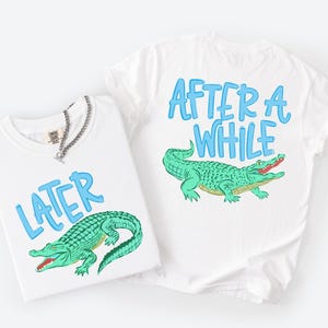 May include: Two white t-shirts with blue text and a green alligator graphic. One shirt says "LATER" and the other says "AFTER A WHILE". A silver chain necklace is on one shirt.