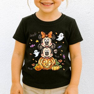 Puede incluir: Camiseta negra con un dise&ntilde;o de Halloween. El dise&ntilde;o muestra a los personajes de Minnie y Mickey Mouse apilados sobre una calabaza, rodeados de fantasmas, murci&eacute;lagos y caramelos. Los personajes llevan disfraces de Halloween.