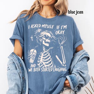 May include: Blue jean-colored t-shirt featuring a skeleton graphic wearing sunglasses and holding a drink. The text on the shirt reads, "I asked myself if I'm okay, and we both started laughing."