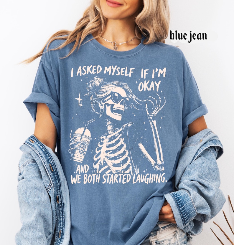 May include: Blue jean-colored t-shirt featuring a skeleton graphic wearing sunglasses and holding a drink. The text on the shirt reads, "I asked myself if I'm okay, and we both started laughing."