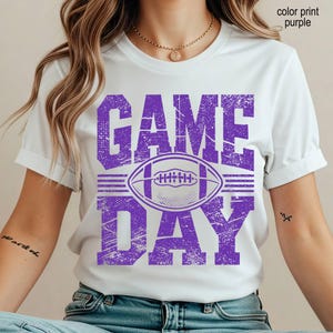May include: A white short-sleeved t-shirt featuring a distressed purple graphic print on the front. The design includes the text "GAME DAY" in large block letters with a football illustration centered between the words. The print has a textured look.