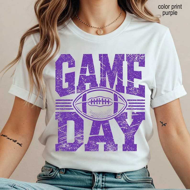 May include: A white short-sleeved t-shirt featuring a distressed purple graphic print on the front. The design includes the text "GAME DAY" in large block letters with a football illustration centered between the words. The print has a textured look.