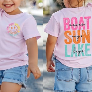 May include: A light pink t-shirt featuring the text "Boat Waves Lake Days" and a smiling sun graphic on the front. The back of the shirt displays "Boat Waves Sun Rays Lake Days" in colourful block letters. The child is wearing blue denim shorts.