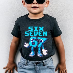 May include: Dark gray t-shirt with the words "SIX SEVEN" in pixelated blue letters. Below, the numbers "67" are depicted as cartoon characters with blue bodies, white gloves, and blue shoes. The shirt is worn by a child.