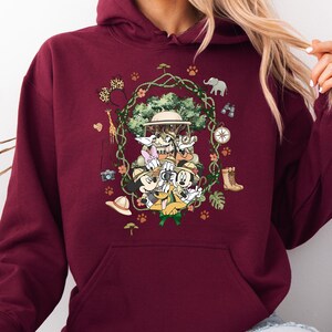 Peut inclure: Sweat &agrave; capuche bordeaux avec un motif sur le th&egrave;me du safari. Le design comprend Mickey Mouse, Minnie Mouse, Donald Duck et Dingo en tenue de safari, entour&eacute;s d'&eacute;l&eacute;ments de la jungle comme une girafe, un &eacute;l&eacute;phant et une boussole.