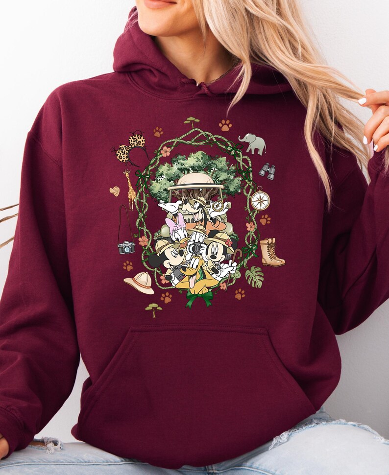 Peut inclure: Sweat &agrave; capuche bordeaux avec un motif sur le th&egrave;me du safari. Le design comprend Mickey Mouse, Minnie Mouse, Donald Duck et Dingo en tenue de safari, entour&eacute;s d'&eacute;l&eacute;ments de la jungle comme une girafe, un &eacute;l&eacute;phant et une boussole.