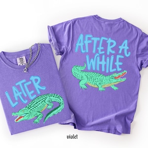 May include: Two violet t-shirts with a green alligator graphic. One shirt says "LATER" and the other says "AFTER A WHILE" in light blue. A silver chain necklace is on the first shirt.