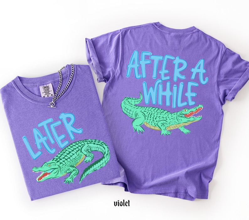May include: Two violet t-shirts with a green alligator graphic. One shirt says "LATER" and the other says "AFTER A WHILE" in light blue. A silver chain necklace is on the first shirt.