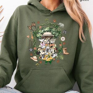 Peut inclure: Sweat &agrave; capuche vert militaire avec un motif de safari repr&eacute;sentant Mickey, Donald, Dingo et Daisy. Le motif comprend une jeep de safari, une boussole, des jumelles et d'autres &eacute;l&eacute;ments de safari. Le texte "military green" est visible.