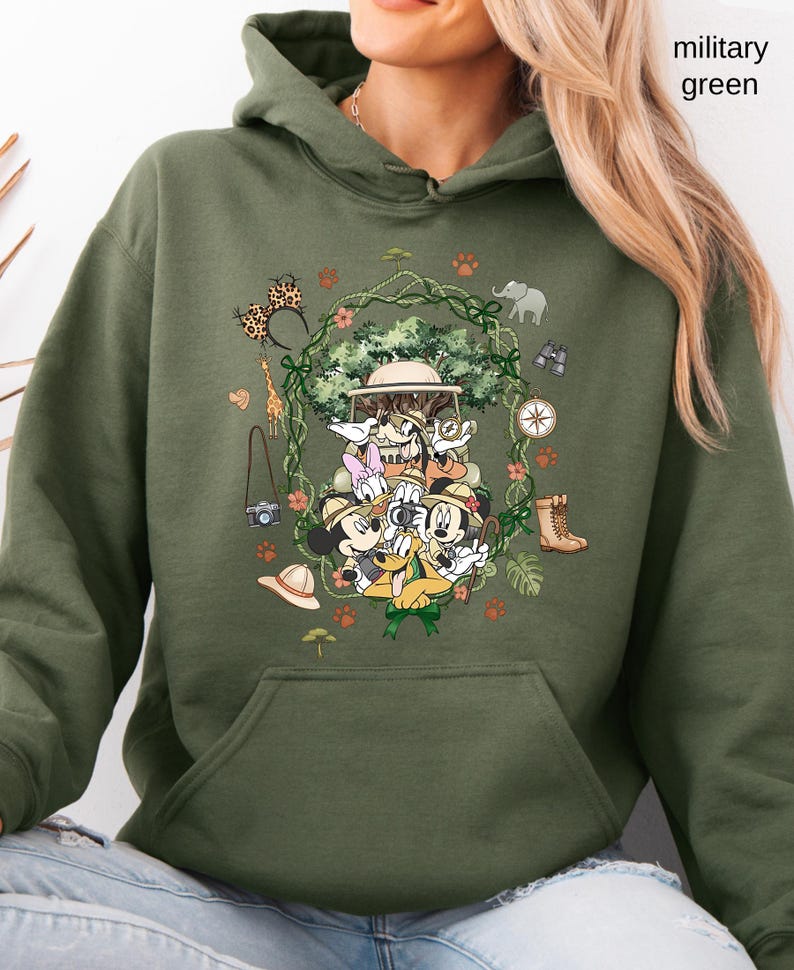 Peut inclure: Sweat &agrave; capuche vert militaire avec un motif de safari repr&eacute;sentant Mickey, Donald, Dingo et Daisy. Le motif comprend une jeep de safari, une boussole, des jumelles et d'autres &eacute;l&eacute;ments de safari. Le texte "military green" est visible.