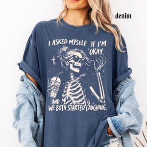 May include: Denim blue t-shirt with a skeleton graphic wearing sunglasses and holding a drink. The text on the shirt reads, "I asked myself if I'm okay, and we both started laughing."