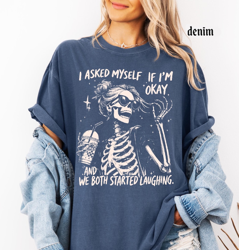 May include: Denim blue t-shirt with a skeleton graphic wearing sunglasses and holding a drink. The text on the shirt reads, "I asked myself if I'm okay, and we both started laughing."