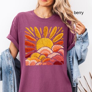 May include: Berry-colored t-shirt featuring an embroidered design of a yellow sun with orange and red rays and clouds. The shirt has rolled-up sleeves and a relaxed fit. The word "berry" is printed on the upper right side of the image.
