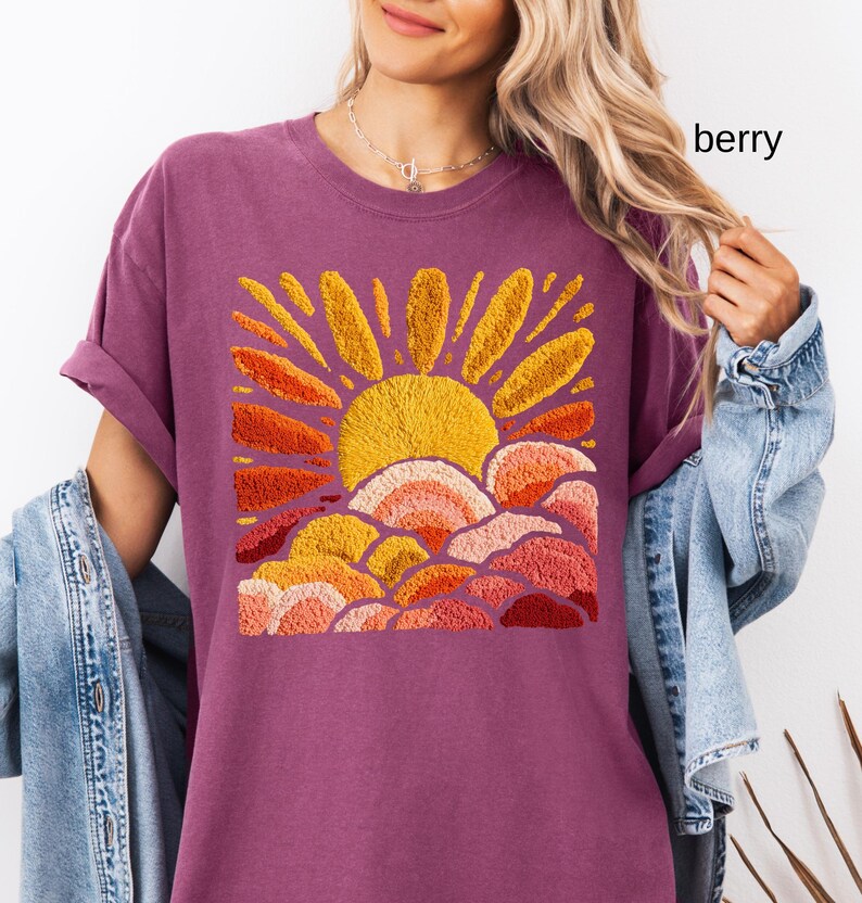 May include: Berry-colored t-shirt featuring an embroidered design of a yellow sun with orange and red rays and clouds. The shirt has rolled-up sleeves and a relaxed fit. The word "berry" is printed on the upper right side of the image.