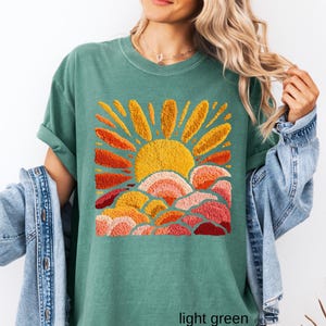 May include: Light green t-shirt featuring an embroidered design of a sun with orange and yellow rays above layered clouds in shades of orange and pink. The text "light green" is at the bottom of the shirt.