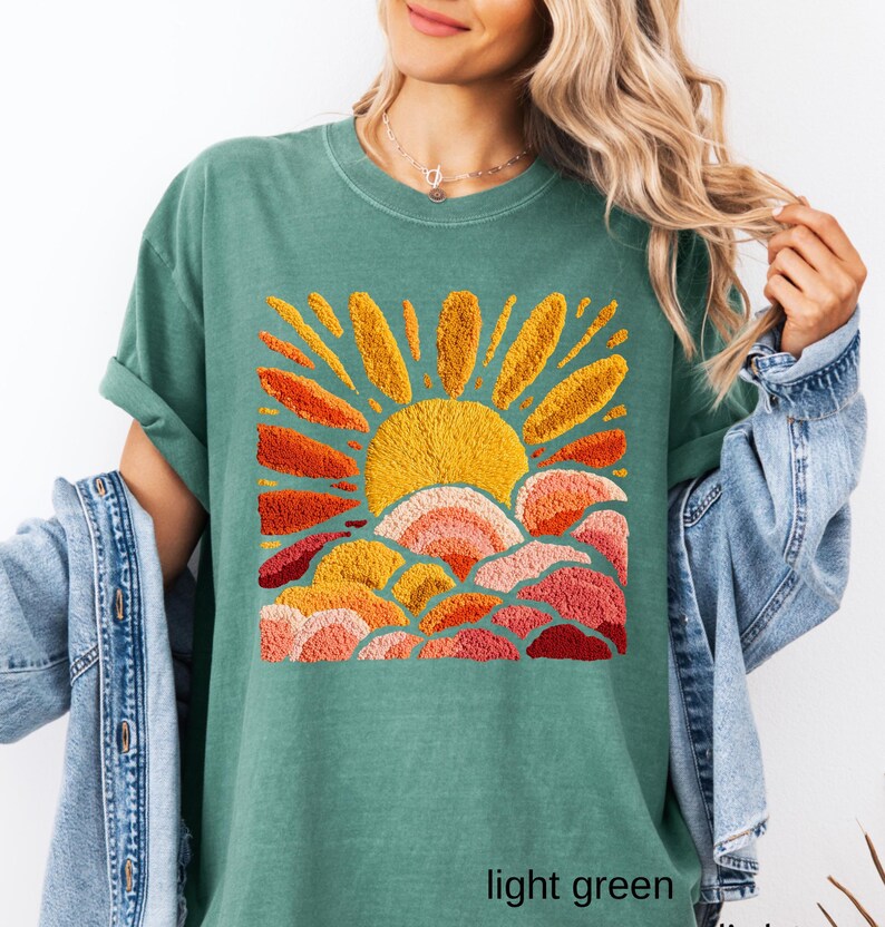May include: Light green t-shirt featuring an embroidered design of a sun with orange and yellow rays above layered clouds in shades of orange and pink. The text "light green" is at the bottom of the shirt.