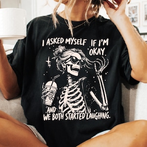 May include: Black t-shirt featuring a skeleton wearing sunglasses and holding a drink. The graphic includes the text "I asked myself if I'm okay, and we both started laughing."