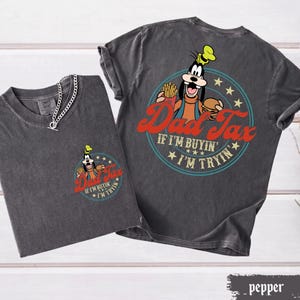 May include: Two charcoal gray t-shirts with the text "Dad Tax" and a cartoon image of Goofy. The front of one shirt has a small graphic, while the other has a larger graphic on the back. The text "If I'm buyin', I'm tryin'" is also included.