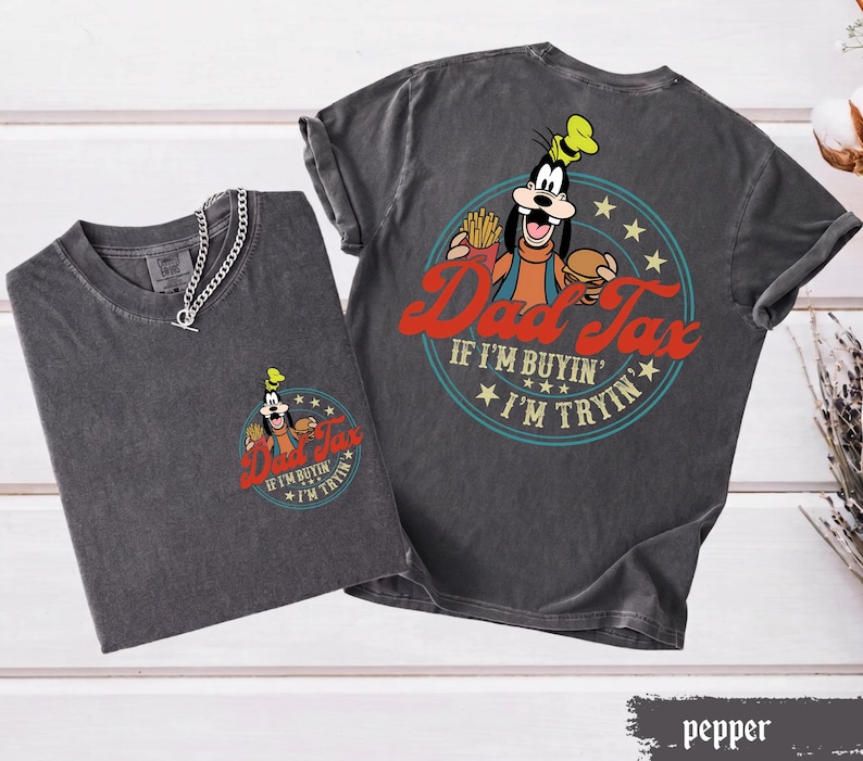 May include: Two charcoal gray t-shirts with the text "Dad Tax" and a cartoon image of Goofy. The front of one shirt has a small graphic, while the other has a larger graphic on the back. The text "If I'm buyin', I'm tryin'" is also included.