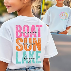 May include: White t-shirt featuring the words "BOAT waves SUN rays LAKE days" in pink, orange, and teal. The shirt also has a smaller design on the front with a smiley face and the words "BOAT WAVES LAKE DAYS".