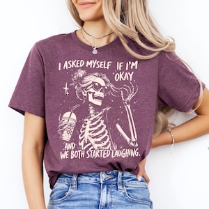 May include: A maroon t-shirt featuring a skeleton graphic wearing sunglasses and holding a drink. The text on the shirt reads, "I ASKED MYSELF IF I'M OKAY. AND WE BOTH STARTED LAUGHING."