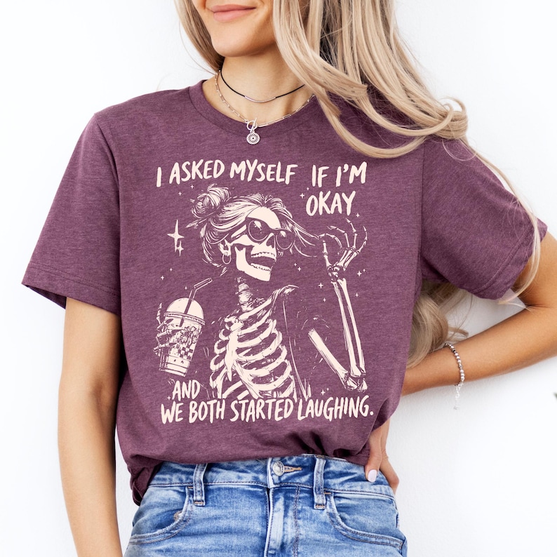 May include: A maroon t-shirt featuring a skeleton graphic wearing sunglasses and holding a drink. The text on the shirt reads, "I ASKED MYSELF IF I'M OKAY. AND WE BOTH STARTED LAUGHING."