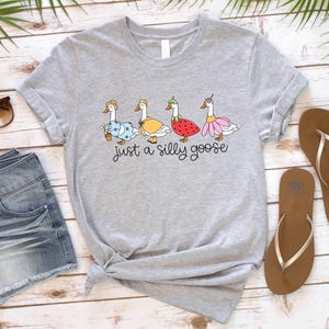 May include: A light gray t-shirt featuring a whimsical illustration of four geese, each adorned with unique outfits and accessories. The text "just a silly goose" is written in a playful font below the geese. The shirt is styled with rolled-up sleeves.