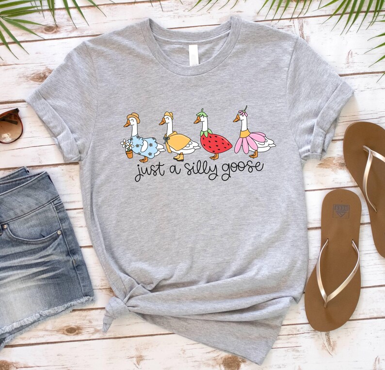 May include: A light gray t-shirt featuring a whimsical illustration of four geese, each adorned with unique outfits and accessories. The text "just a silly goose" is written in a playful font below the geese. The shirt is styled with rolled-up sleeves.
